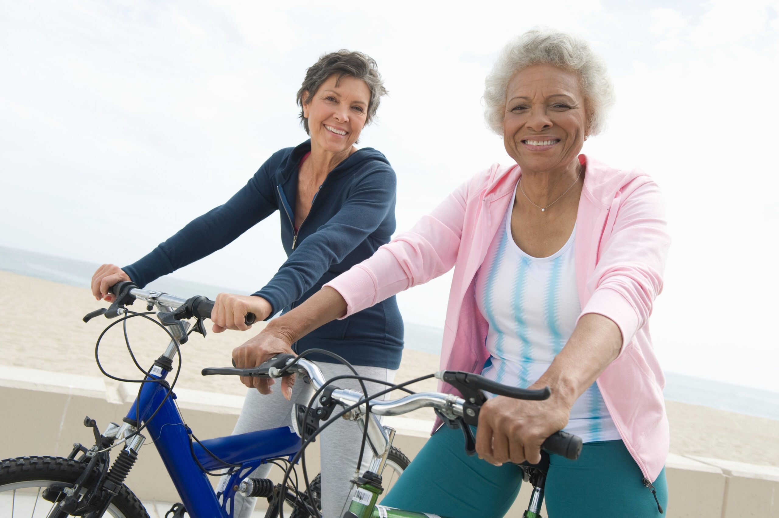 Two active senior women with bright smiles enjoying a bike ride by the beach, living life to the fullest without pain.
